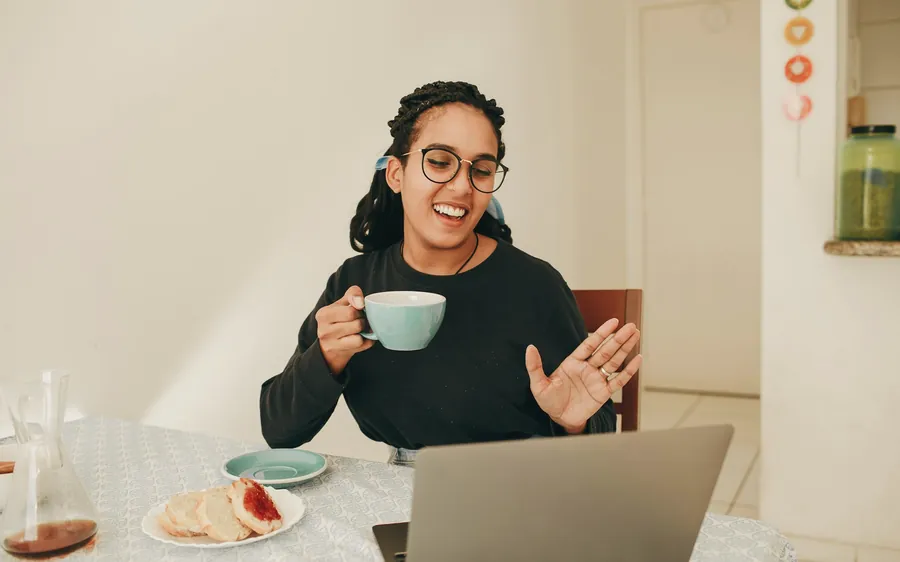 A woman in glasses laughs as she talks on a video call at her kitchen table, holding a mug of tea.