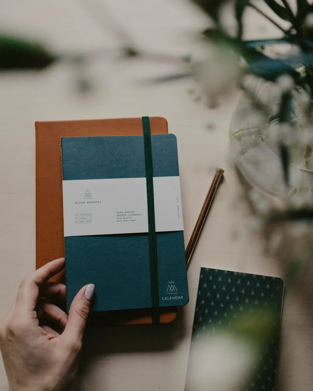 Hands hold a dark green notebook and an orange one, with a pencil beside them.