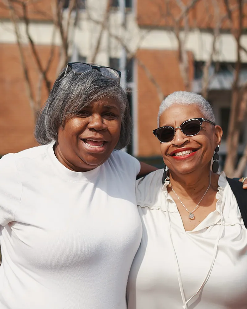 Two grey-haired friends stand arm in arm outside, both laughing.