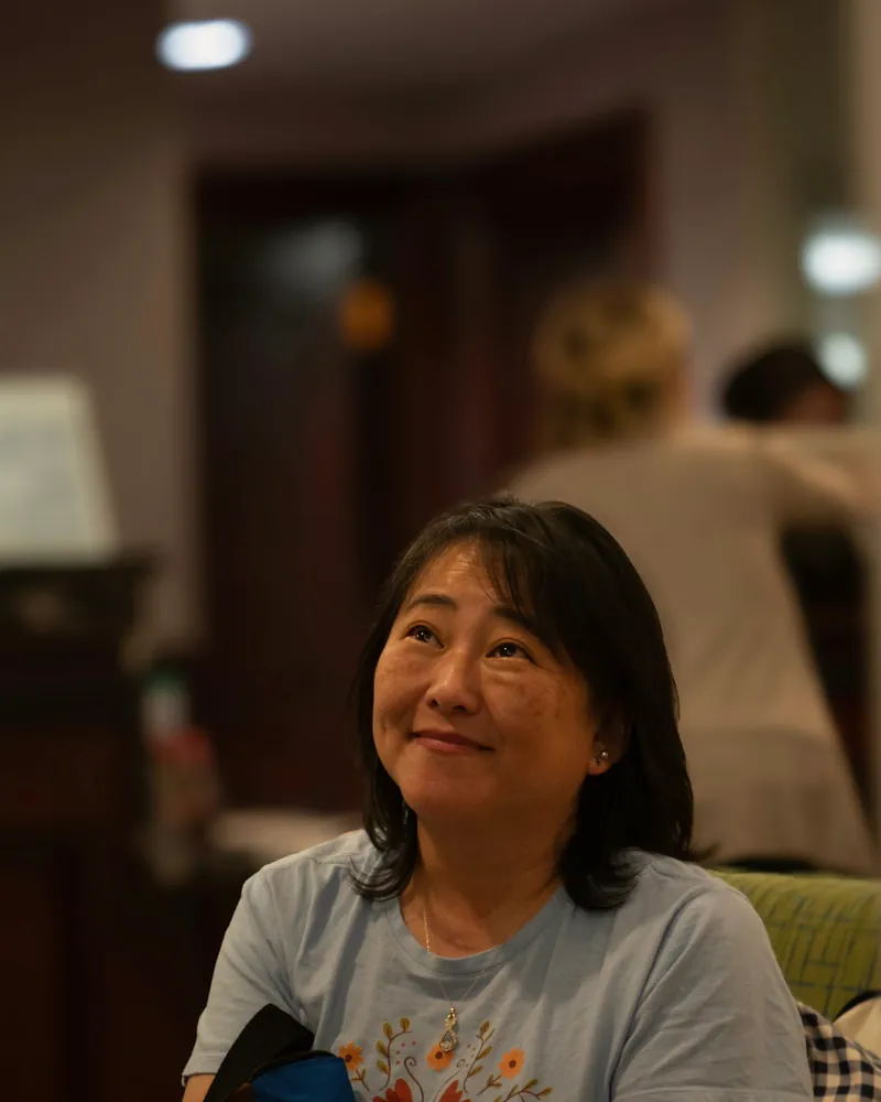 A woman in her fifties sits in her living room, smiling as she listens to someone.