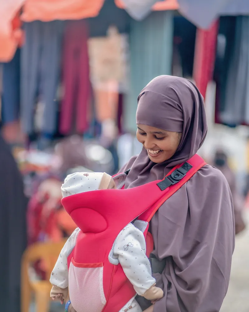 A mother in a hijab smiles down at her baby, who is snug in a red carrier on her front.