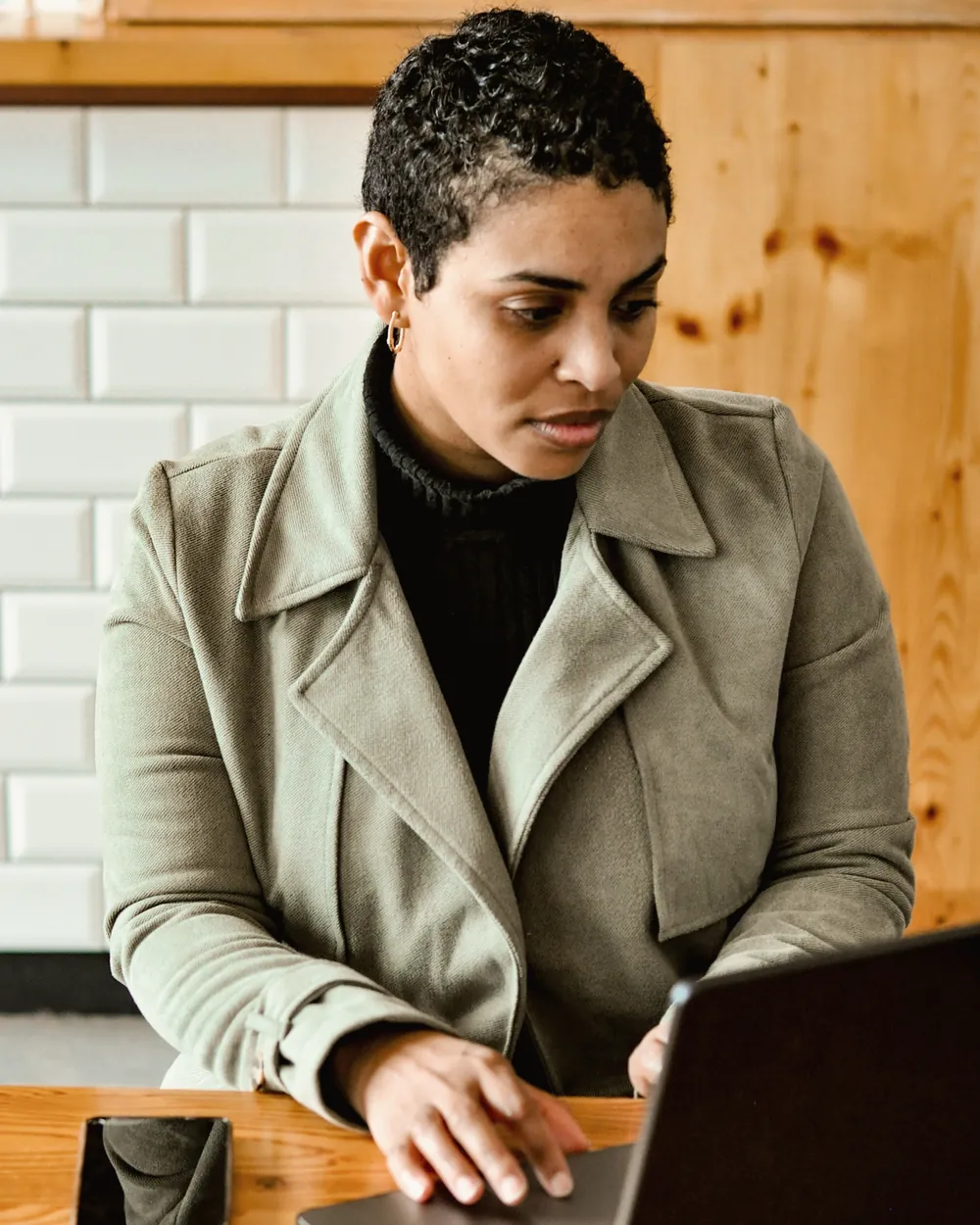 A woman in a green coat types on her laptop at a wooden table, her phone beside her.