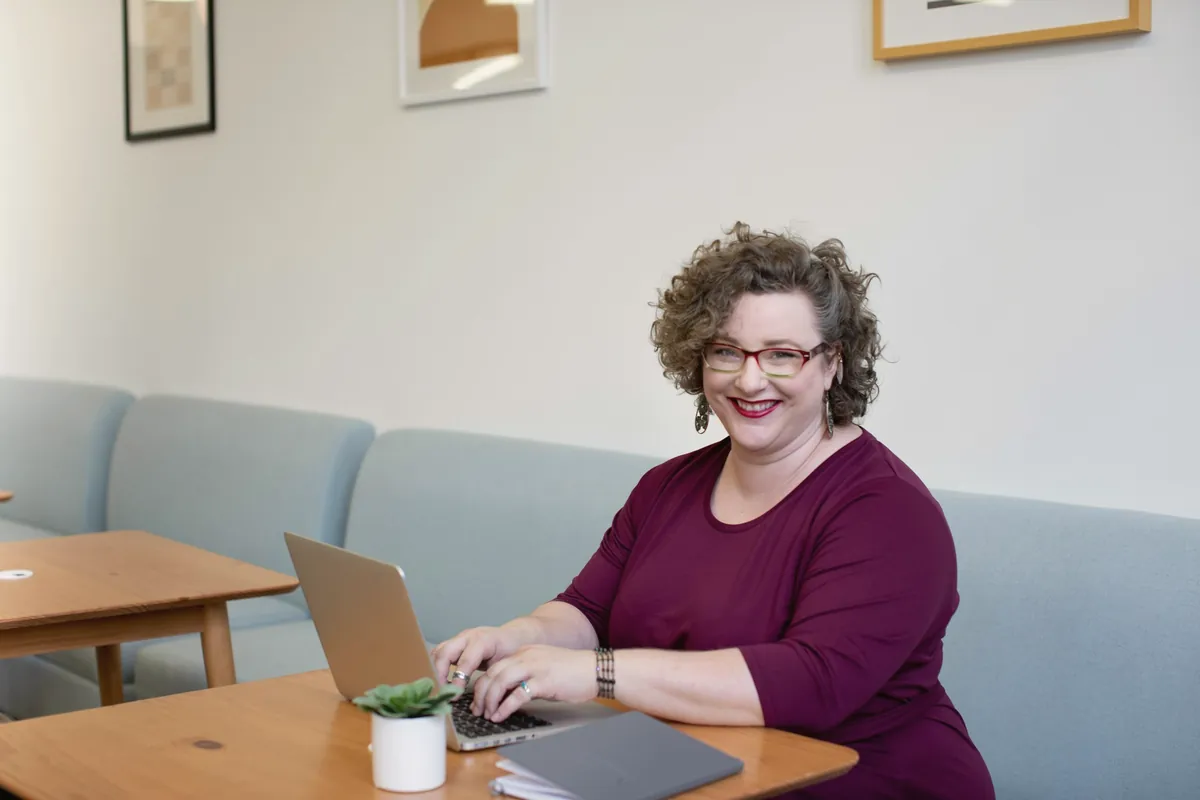 A woman with curly hair and red glasses works on her laptop in a bright office.