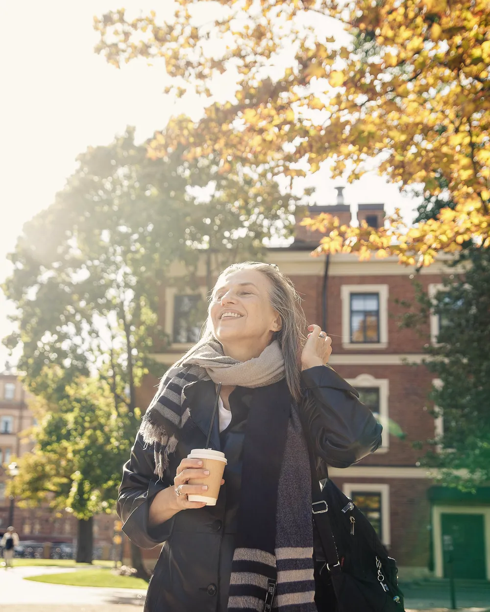 A woman with a scarf and a takeaway coffee walks under autumn trees, smiling.