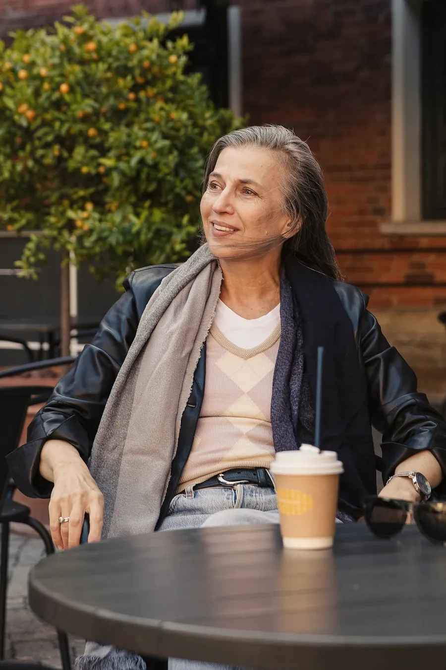 A woman with greying hair sits at a café table outside with a takeaway coffee, looking up and smiling.