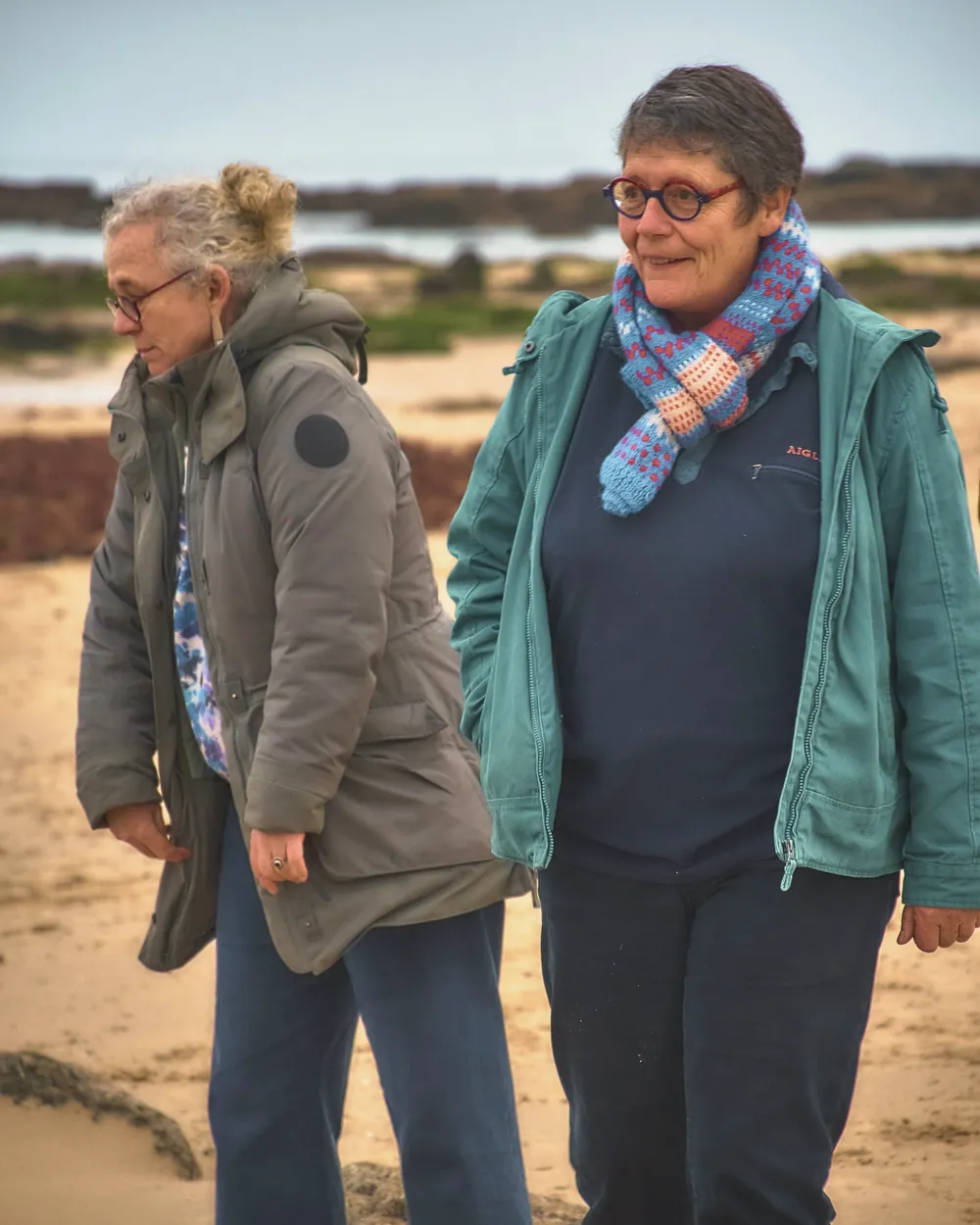 Two women in fleeces and scarves chat as they walk along a sandy beach.