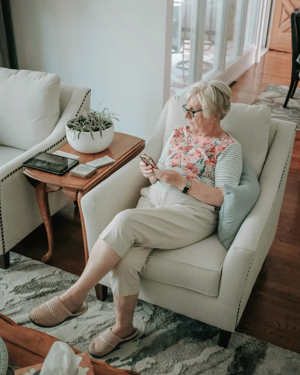 A woman with short silver hair sits in an armchair by the window, reading on her phone.