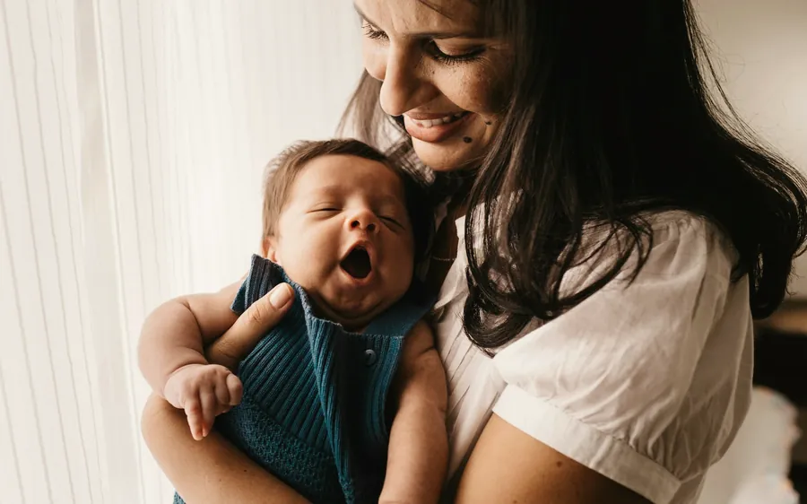 A mother smiles down at the yawning newborn in her arms, by a bright window.
