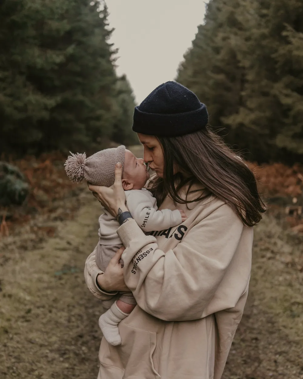 A mother in a woolly hat holds her baby close on a woodland path.